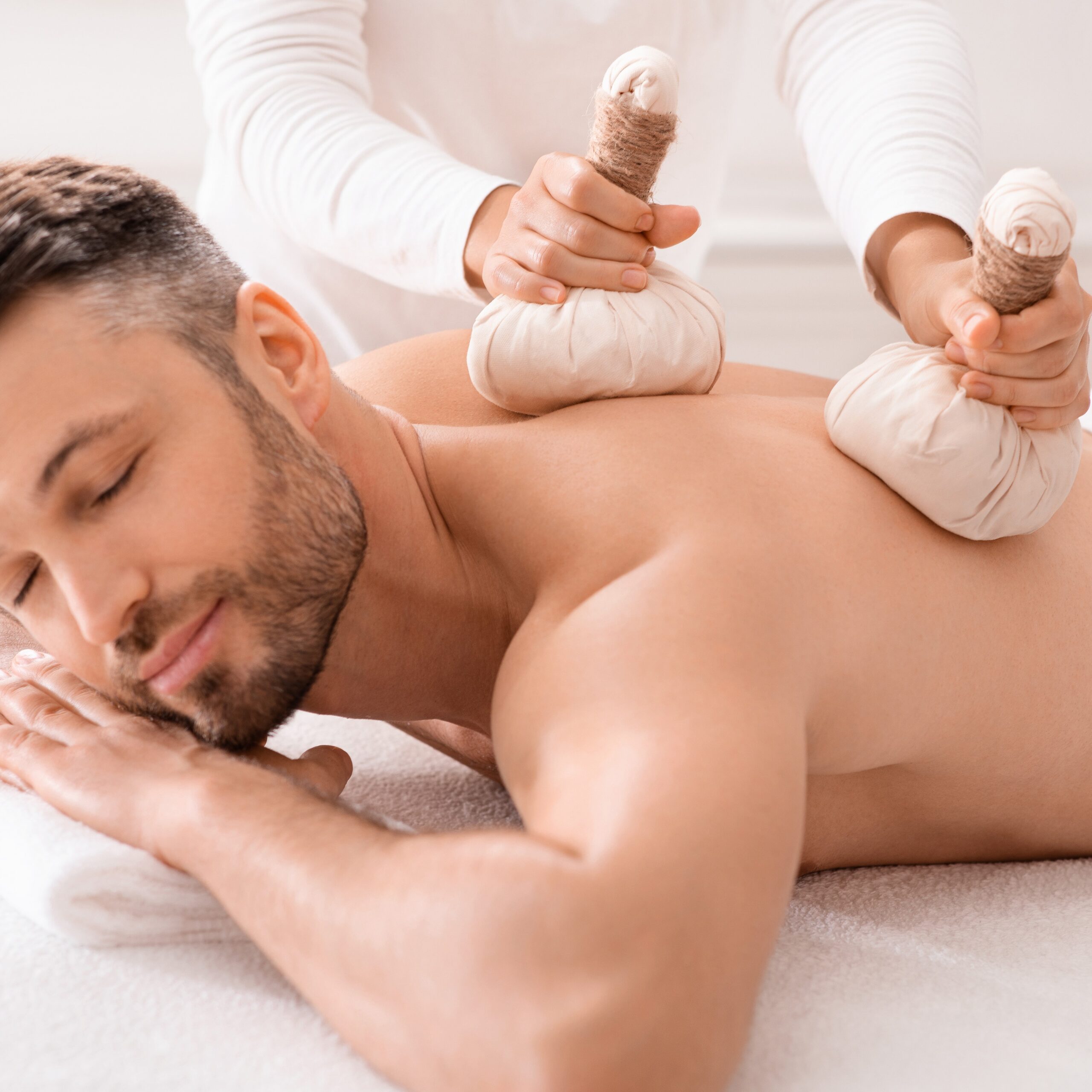 Herbal massage concept. Unrecognizable spa attendant making body massage for handsome man with herbal compress balls, luxury spa interior. Man enjoying herbal treatment at massage salon